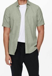 Uomo che indossa una camicia a maniche corte verde chiaro con bottoni sopra una maglietta bianca e pantaloni neri, con una mano in tasca e un braccialetto d'argento al polso.