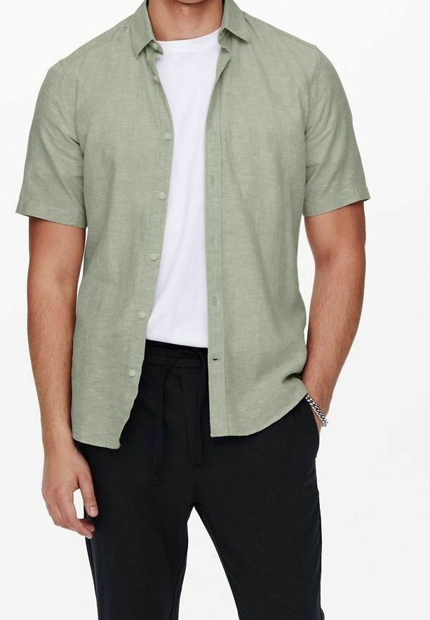 Uomo che indossa una camicia a maniche corte verde chiaro con bottoni sopra una maglietta bianca e pantaloni neri, con una mano in tasca e un braccialetto d'argento al polso.