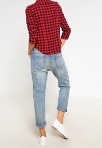 Röd och svart rutigt knäppskjorta, bärs med ljusblå rullade jeans och vita sneakers. Skjortan har långa ärmar och en rundad nederkant.