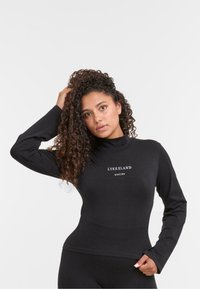 Femeie cu păr creț, îmbrăcată într-un top negru cu mânecă lungă pe care scrie "LYKKELAND atelier", pozând cu o mână pe cap și cealaltă pe șold.