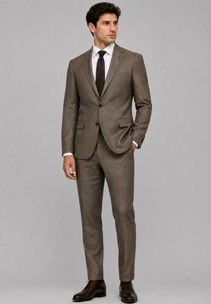 Homme portant un costume marron deux pièces avec une chemise blanche et une cravate noire, debout avec une main dans la poche, regardant sur le côté sur un fond gris.