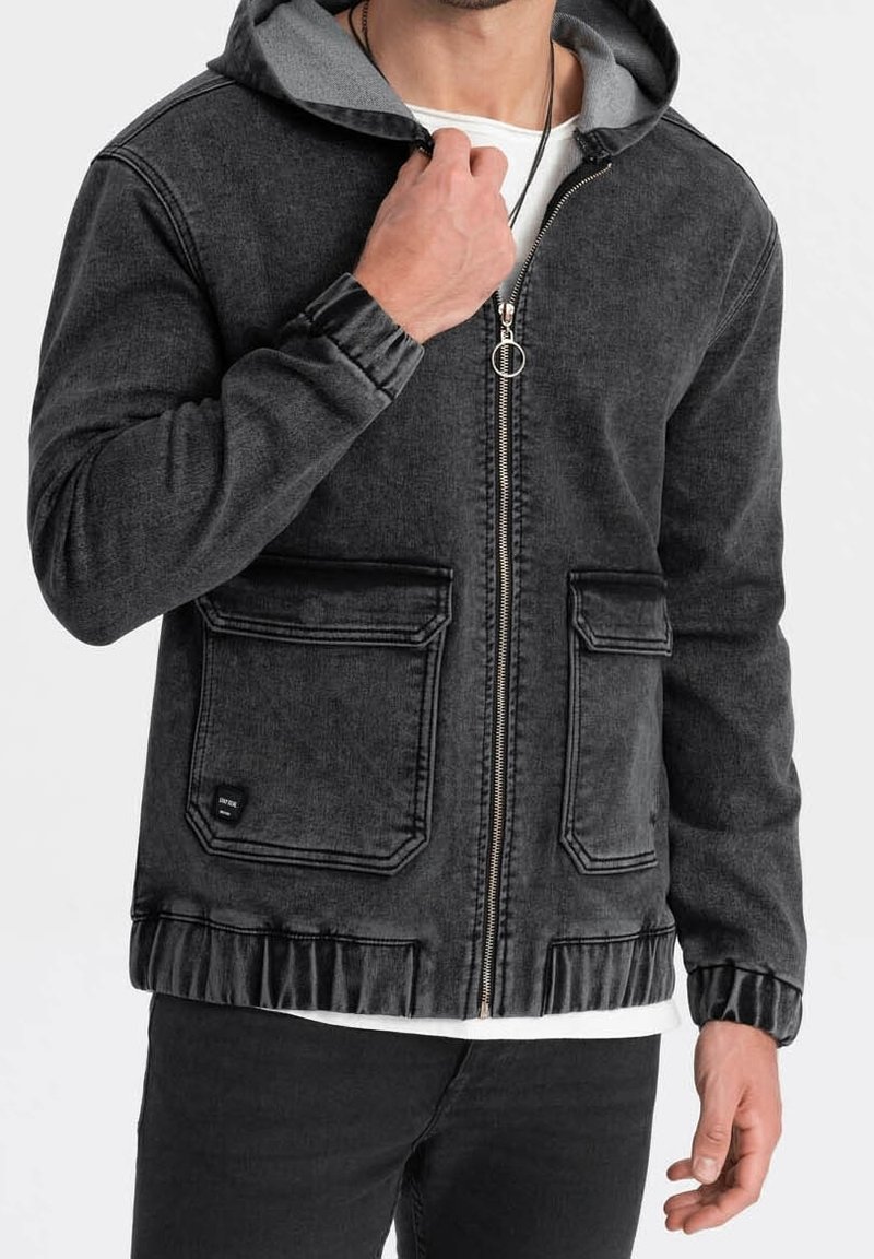 Homme portant une veste noire à capuche avec fermeture éclair, deux poches sur le devant, des poignets élastiques, et une chemise blanche en dessous.