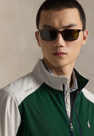 Homme portant des lunettes de soleil écaille de tortue et une veste zippée Ralph Lauren verte et blanche par-dessus une chemise rayée, faisant face à l'avant.