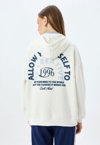 Felpa con cappuccio color crema realizzata in tessuto morbido, con grande testo blu navy sulla schiena, che riporta "1996" e frasi motivazionali.