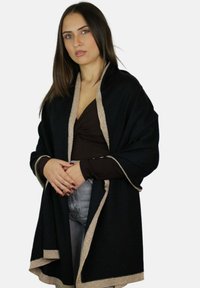 Jeune femme aux longs cheveux bruns portant un châle noir à bordure beige sur un haut marron et un jean gris, debout les mains jointes.