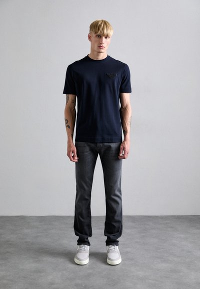 T-shirt marine a maniche corte con dettaglio logo testurizzato sul petto, abbinato a jeans grigi e sneakers grigie chiare.