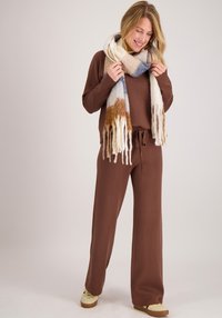 Bruine gebreide trui en bijpassende joggingbroek, gecombineerd met een dikke, meerkleurige sjaal met blauw, crème en roestkleurige patronen.