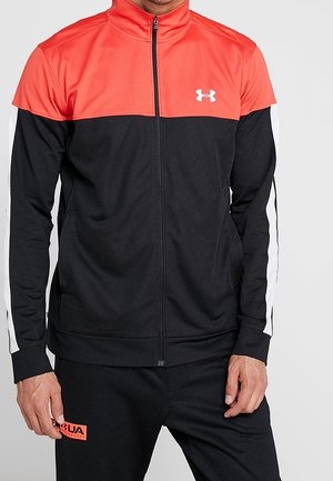 Veste de survêtement zippée noire et rouge avec des rayures blanches, fabriquée en tissu doux et texturé. Présente un col haut et un logo sur la poitrine.