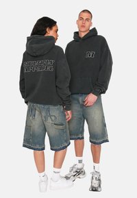 Tmavě šedá mikina s kapucí s nápisem "MULTIPLY APPAREL" na zádech, doplněná o vybledlé modré denimové šortky s dekoračním prošitím a kapsami.