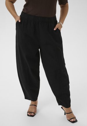 Donna che indossa pantaloni neri larghi con vita elasticizzata, top marrone e tacchi neri aperti sulle dita, in piedi con le mani nelle tasche su sfondo bianco.