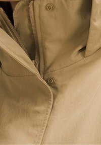 Gros plan d'une veste beige montrant une fermeture éclair partiellement ouverte et des boutons-pression sur un tissu lisse.