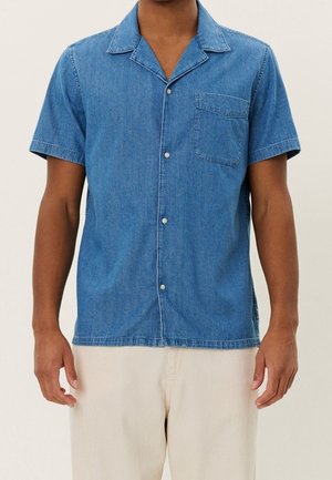 Man wearing a short-sleeve blue denim button-up shirt with a chest pocket and light-colored pants standing against a plain background.