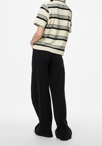 En person står med ryggen mot kameran, iklädd en randig kräm- och svart skjorta samt vida svarta byxor mot en neutral bakgrund.