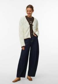 Cardigan en tricot crème avec motif en losange ; bordure brun foncé. Associé à un t-shirt graphique marron et un jean large en denim foncé. Ballerines marron.