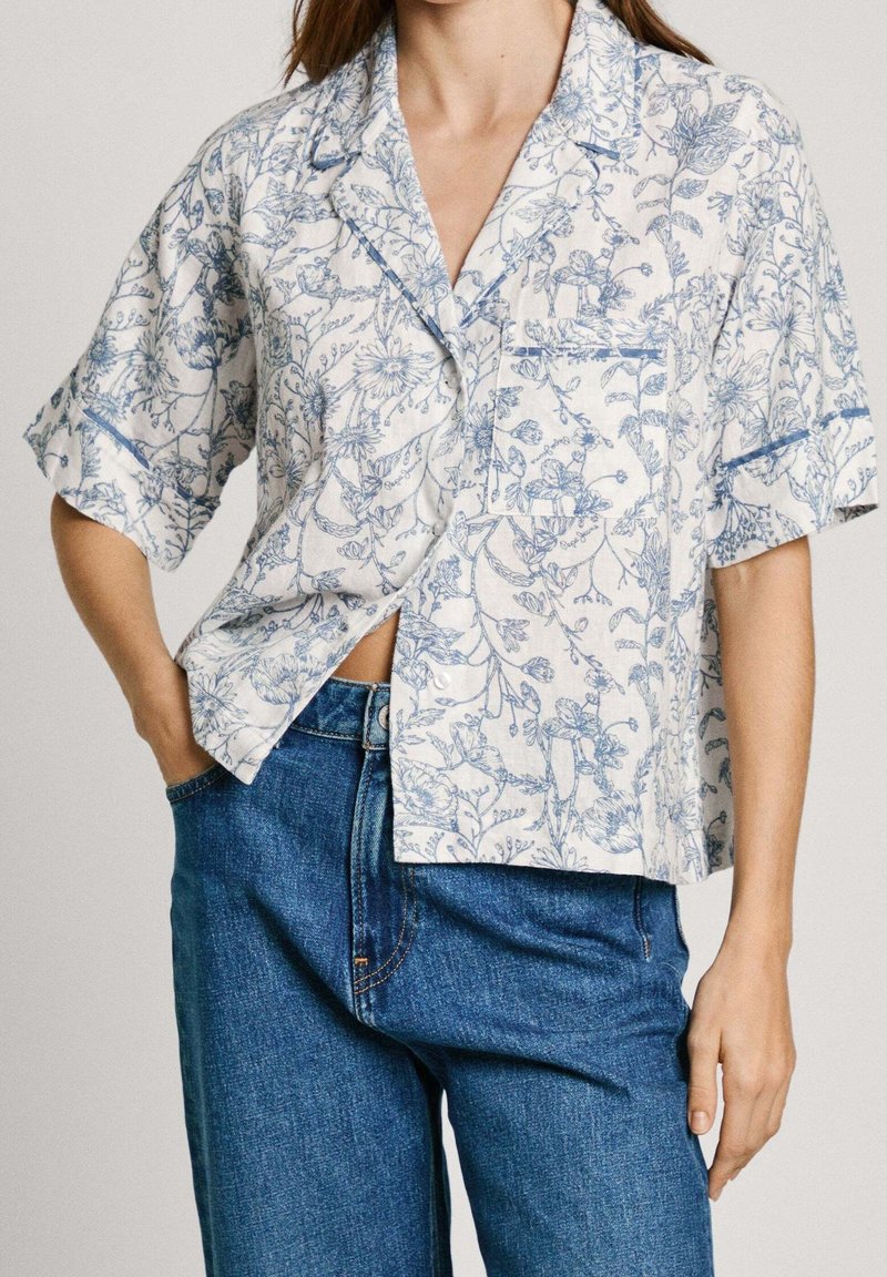 Femme portant une chemise blanche ample à manches courtes avec un imprimé floral bleu et un jean bleu, une main dans la poche, debout devant un fond uni.