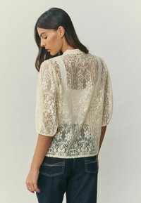 Mežģīņu blūze kremkrāsā ar pūderētām piedurknēm, kas izrotātas ar ziedu un ģeometriskiem rakstiem, caurspīdīgu tekstūru un apaļām apakšmalas dizainu.