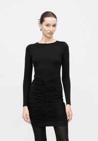 Γυναίκα με μαλλιά μαζεμένα προς τα πίσω, φορώντας εφαρμοστό μαύρο μίνι φόρεμα με ρυτιδώσεις και μαύρες καλσόν, μπροστά σε απλό λευκό φόντο.