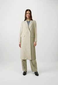 Casaco bege de abotoamento duplo com botões grandes, bolsos laterais e tecido texturizado; combinado com calças cargo claras e sapatos pretos.