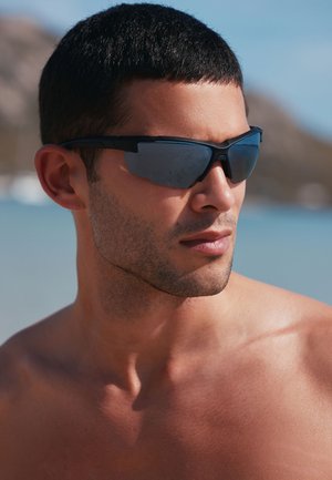 Black sports sunglasses with mirrored lenses, angular design, and sturdy frame. Close-up of face; bare skin and short dark hair visible.