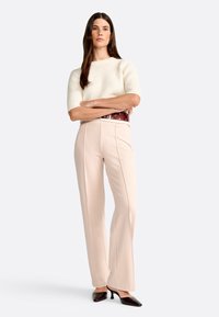 Maglione corto a maniche corte in misto crema, pantaloni a vita alta di un tenue rosa pallido con pieghe frontali e cintura bordeaux. Décolleté neri a punta.