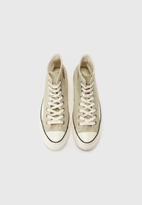 Hoch-top Sneaker aus hellbeigem Canvas mit weißen Schnürsenkeln und Gummikappen an den Zehen. Merkmale sind braune Akzente und schwarze Paspelierung entlang der Sohle.