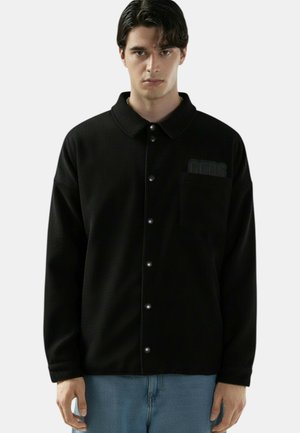 Giovane uomo che indossa una giacca nera con bottoni e taschino sul petto, abbinata a jeans azzurri, in piedi davanti a uno sfondo chiaro e neutro.