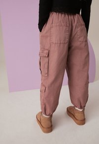 Dulkiai rožinės medvilnės cargo kelnės su elastiniais kulkšniais, dviem cargo kišenėmis ir laisvu siluetu, derinamos su rudais zomšiniais kulkšnies batais.