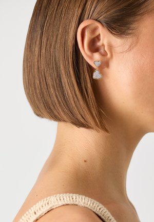 Gold heart-shaped earrings with sparkling silver accents, featuring a stacked design. The earrings hang from a post style, complementing short hair.