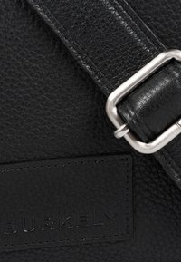 Borsa in pelle nera con superficie testurizzata, dotata di una fibbia in metallo argentato e branding in rilievo su una toppa rettangolare.