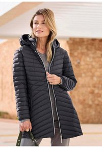 Zwarte pufferjas met een ritssluiting aan de voorkant, een capuchon en chevron-quilting. Heeft witte bies en een relaxed model, gecombineerd met een grijze top.