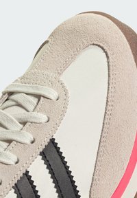 Béžová a bílá sportovní obuv s semišovými a síťovinovými materiály, černými vroubkovanými pruhy, bílými tkaničkami a růžovou gumovou podrážkou.