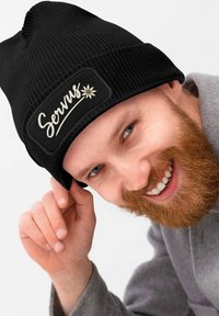 Black knitted beanie with a rectangular black patch featuring the word "Servus" and a flower design. The fabric appears textured.