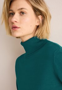 Maglione a collo alto in teal realizzato in materiale liscio e leggero con collo a costine. Presenta maniche lunghe e una silhouette aderente.