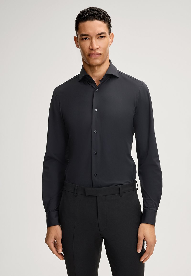 Maglietta nera a maniche lunghe con colletto a punta, chiusura frontale con bottoni e texture liscia. Abbinata a pantaloni neri, senza motivi visibili.