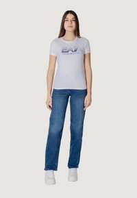 T-shirt bianca a maniche corte con stampa grafica "EA7 EMPORIO ARMANI", abbinata a jeans blu e sneakers bianche. Design semplice e forma aderente.