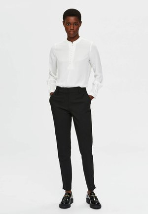 Blusa blanca de manga larga con botones en la parte delantera, combinada con pantalones negros ajustados y mocasines negros con suela gruesa y acentos dorados.