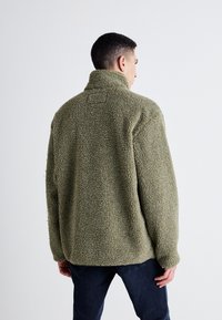 Giacca in pile verde oliva con collo alto, vestibilità comoda e superficie testurizzata. Presenta un'etichetta quadrata sul retro.