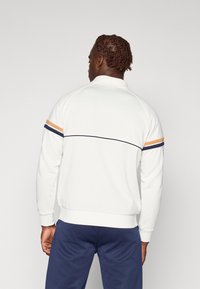 Hvit sweatshirt med høy krage, med blå og oransje striper på skuldrene. Båret med blå bukser, viser en avslappet passform.