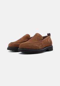 Brown suede slip-on shoes with a rounded toe, featuring contrasting stitching and a black rubber sole. Minimalist design with a back pull tab.