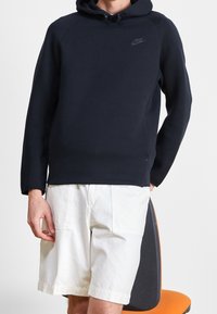 Mörkblå huppare med dragsko, ribbade muddar och en diskret logotyp på bröstet, kombinerad med vita cargoshorts på en sittande figur.