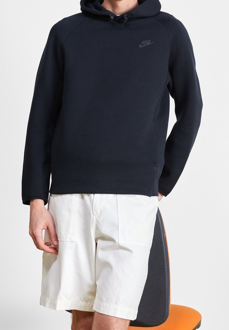 Mörkblå huppare med dragsko, ribbade muddar och en diskret logotyp på bröstet, kombinerad med vita cargoshorts på en sittande figur.