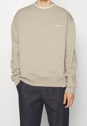 Sweat-shirt beige avec petit texte "LES DEUX" sur la poitrine, porté avec un pantalon gris foncé, mains détendues sur les côtés.