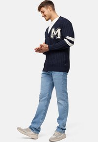Marineblå kabelstrikket cardigan med hvide striber og et fremtrædende hvidt "M" på brystet, sat sammen med lyseblå jeans og beige sneakers.