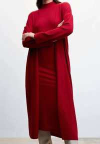 Femme portant une robe rouge à manches longues, longue jusqu'au sol, avec un cardigan assorti à manches longues, debout les bras croisés devant un fond uni.
