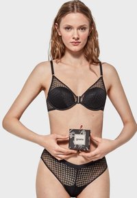 Sujetador negro con una superposición de malla geométrica y copas acolchadas, combinado con bragas de talle alto a juego. La modelo sostiene una caja decorativa negra.
