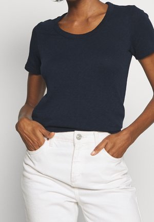 Mujer con camisa de manga corta azul marino metida en pantalones blancos de tiro alto, con las manos en los bolsillos delanteros.