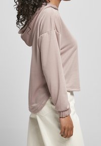 Růžová mikina s kapucí, volného střihu, dlouhými rukávy a ribovanými manžetami. Tkanina vypadá měkce a hladce, s mírně strukturovaným povrchem.