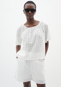 Completo di cotone bianco caratterizzato da una parte superiore ampia con scollatura rotonda e maniche corte, abbinato a pantaloni corti coordinati. Entrambi presentano un motivo a griglia.