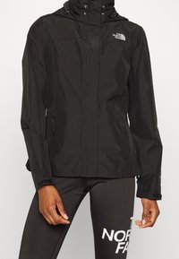 Zwarte waterdichte jas met capuchon, rits en logo op de borst, gedragen met zwarte leggings met grote witte tekst "The North Face" op de dij.