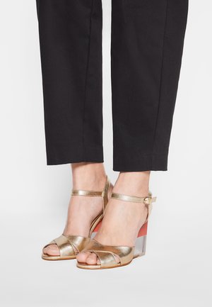 Guldmetalliske sandaler med blokhæle, med gennemsigtig hæl- og ankelrem; krydset remdesign fremhæver foden.